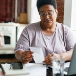 Portrait of puzzled upset serious overweight african american grandma in glasses reading paycheck or utility bill attentively sitting in her kitchen in front of laptop and smartphone